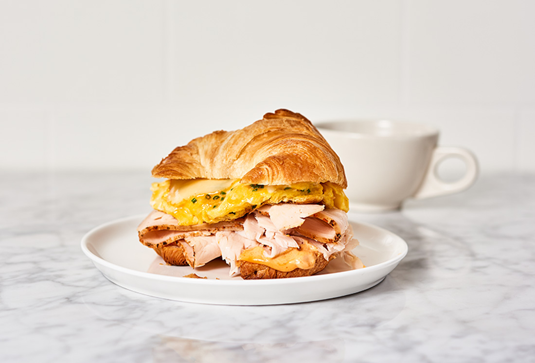 Jennie-O Golden Roasted Turkey Breast Breakfast Sandwich on a plate with a coffee cup behind it.