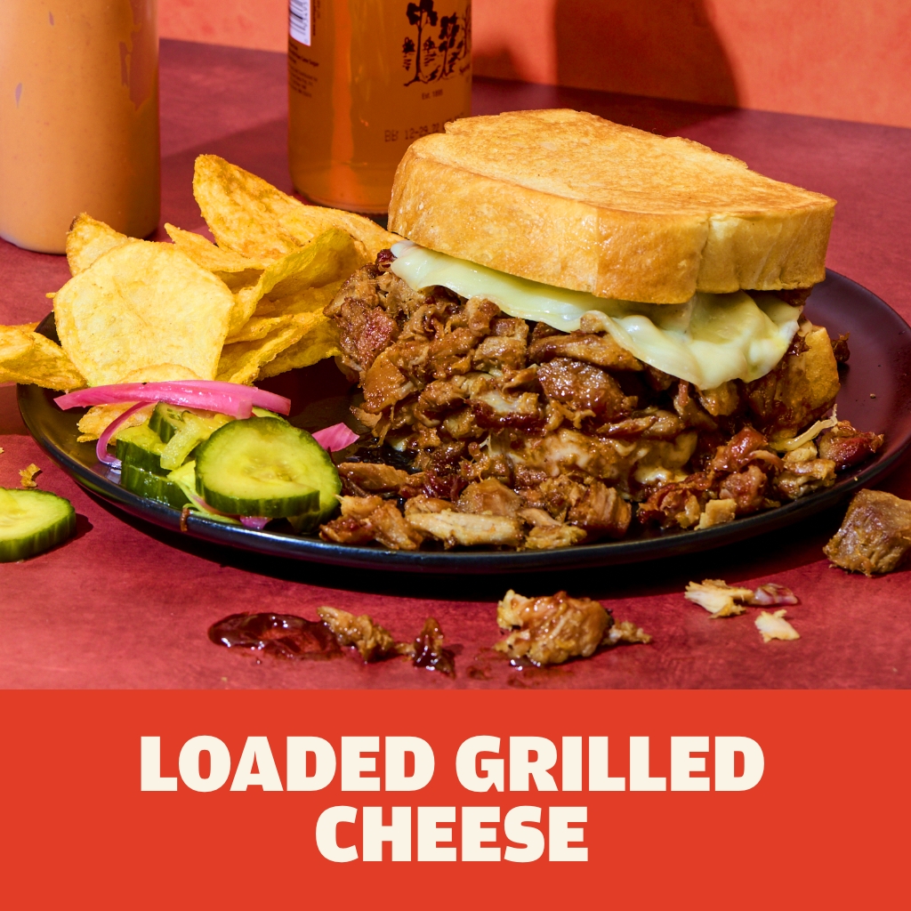 AUSTIN BLUES Hardwood Pulled Pork loaded grilled cheese on Texas toast served on a black plate with kettle chips and pickles.