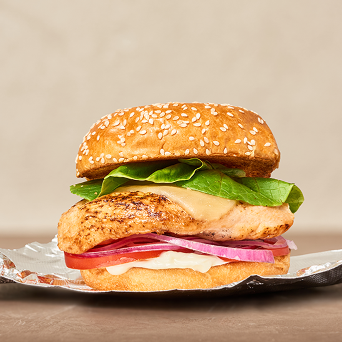 Side view of a sandwich with Fire Braised Chicken Breast on a sesame bun with bacon, lettuce, tomato, red onion and mayo on foil wrapper.