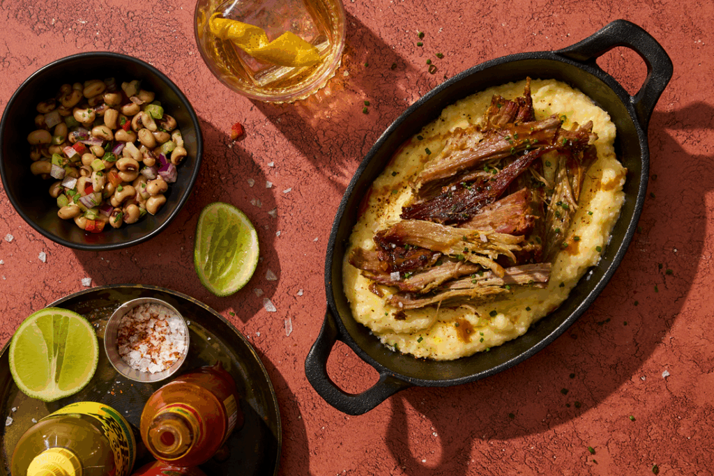 Pork shoulder pork grits in cast iron dish with side of black eyed peas and hot sauces.