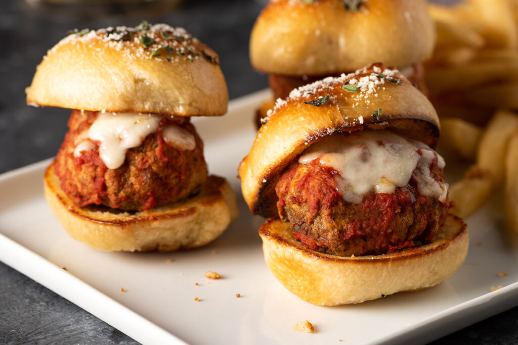 Garlic knot meatball sliders topped with sauce and cheese on a white tray.