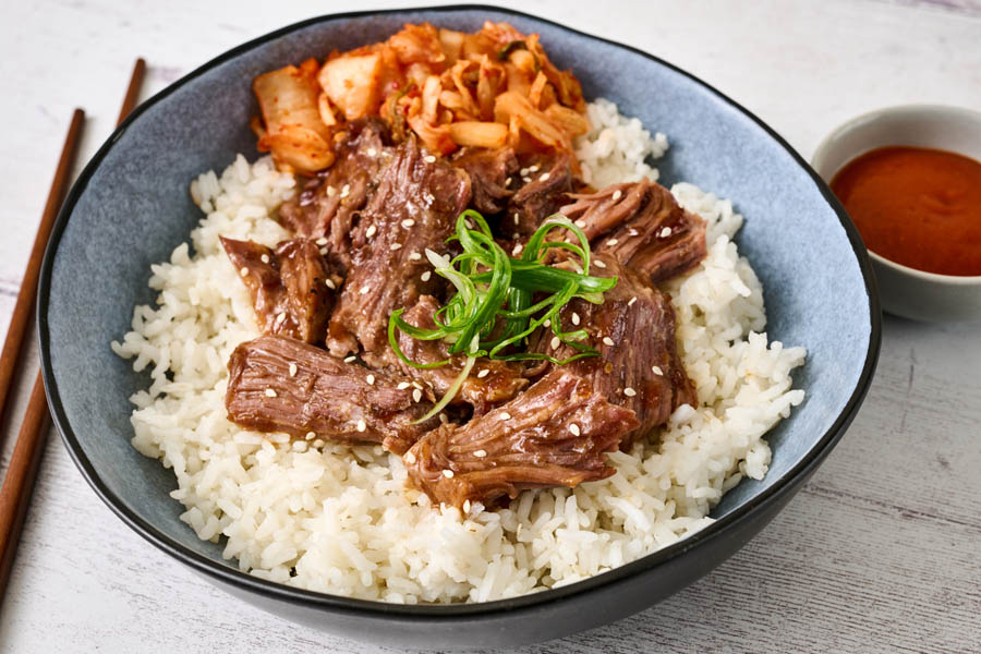 Korean-inspired pot roast beef bowl with rice and garnish, served with a side of sauce
