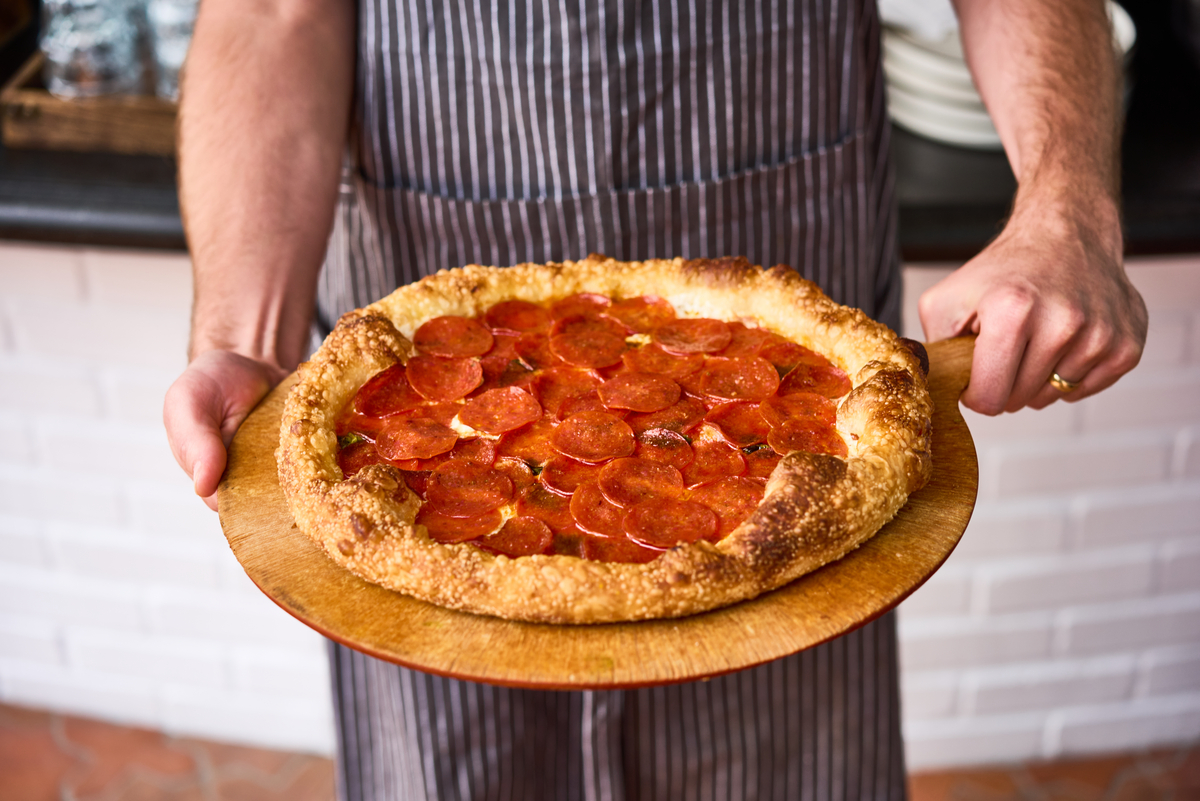 Chef holding a Performance Pepperoni Bold Traditional pepperoni pIzza on a round, wooden pizza peel.